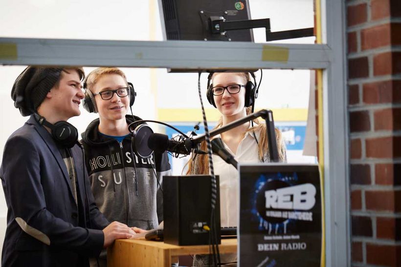 Drei Schüler mit Kopfhörern versammelt um ein Mikrofon