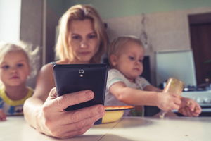 Symbolbild: eine Mutter schaut auf ihr Handy während sie ihre Kinder auf dem Schoß hat
